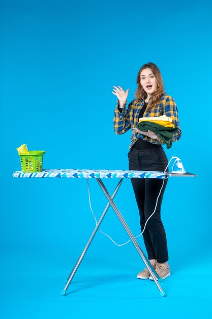 Vorderansicht der jungen Frau, die die gefaltete Kleidung hält, die fünf zeigt und hinter dem Bügelbrett in der Waschküche auf blauem Tisch steht