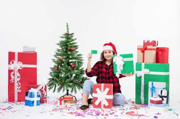 Vorderansicht der jungen Frau, die herum sitzt, präsentiert grüne Bankkarte und Geschenk auf weißer Wand