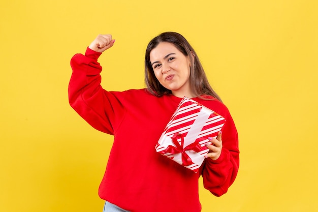 Vorderansicht der jungen Frau, die kleines Geschenk auf gelber Wand hält