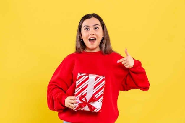 Vorderansicht der jungen Frau, die kleines Geschenk auf gelber Wand hält