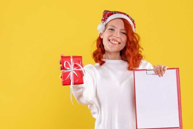 Vorderansicht der jungen Frau, die kleines Weihnachtsgeschenk und Aktennotiz auf gelber Wand hält