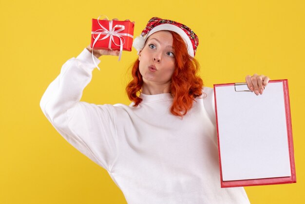 Vorderansicht der jungen Frau, die kleines Weihnachtsgeschenk und Aktennotiz auf gelber Wand hält