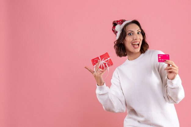 Vorderansicht der jungen Frau, die kleines Weihnachtsgeschenk und Bankkarte auf rosa Wand hält