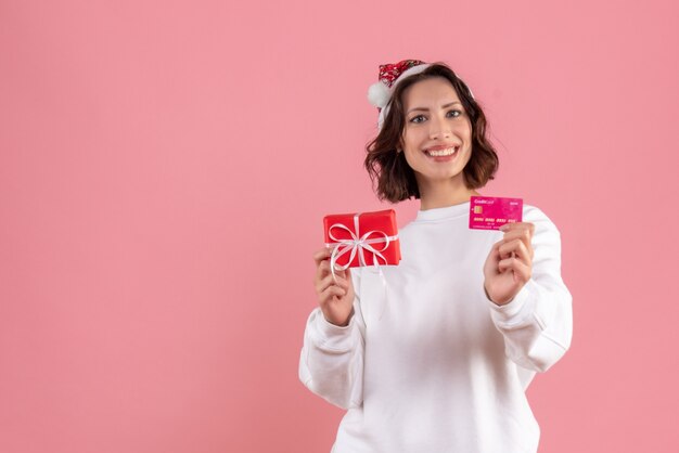 Vorderansicht der jungen Frau, die kleines Weihnachtsgeschenk und Bankkarte auf rosa Wand hält
