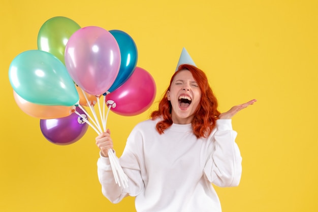 Vorderansicht der jungen Frau, die niedliche bunte Luftballons auf gelber Wand hält