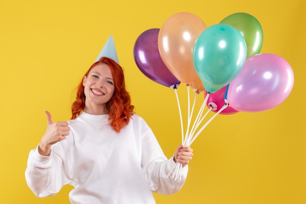 Vorderansicht der jungen Frau, die niedliche bunte Luftballons auf gelber Wand hält