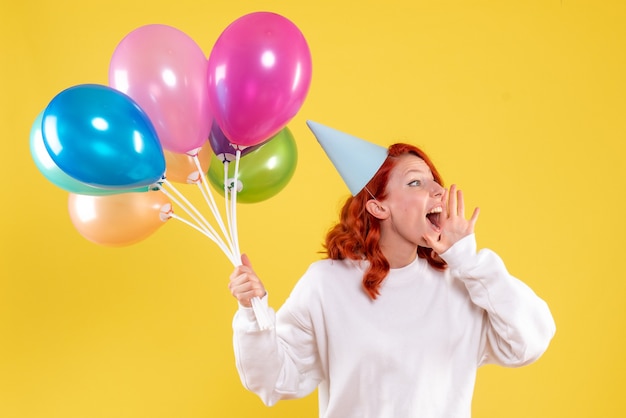 Vorderansicht der jungen Frau, die niedliche bunte Luftballons auf gelber Wand hält