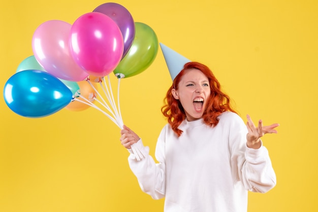 Vorderansicht der jungen Frau, die niedliche bunte Luftballons auf gelber Wand hält