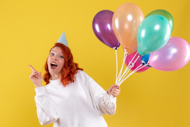 Vorderansicht der jungen Frau, die niedliche bunte Luftballons auf gelber Wand hält