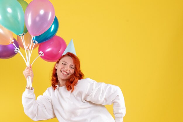 Vorderansicht der jungen Frau, die niedliche bunte Luftballons auf gelber Wand hält