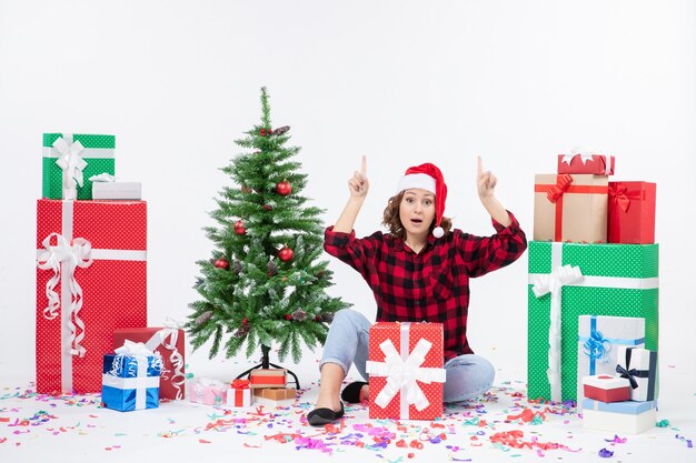 Vorderansicht der jungen Frau, die um Geschenke und kleinen Feiertagsbaum auf weißer Wand sitzt