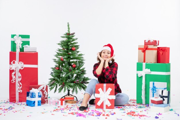 Vorderansicht der jungen Frau, die um Geschenke und kleinen Feiertagsbaum auf weißer Wand sitzt