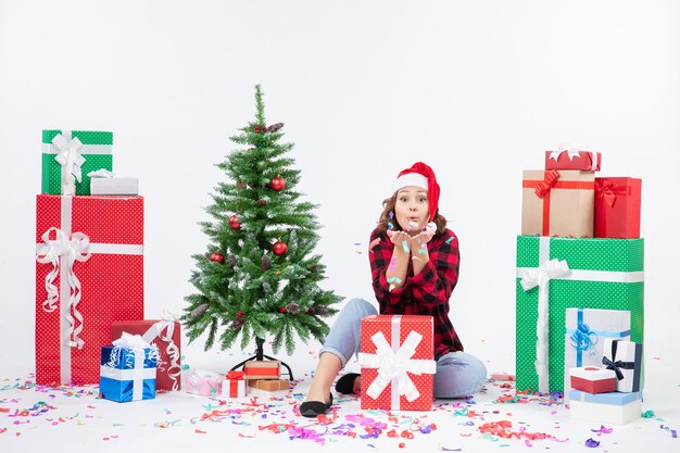 Vorderansicht der jungen Frau, die um Geschenke und kleinen Feiertagsbaum auf weißer Wand sitzt