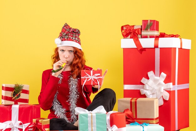 Vorderansicht der jungen Frau, die um verschiedene Weihnachtsgeschenke auf gelber Wand sitzt