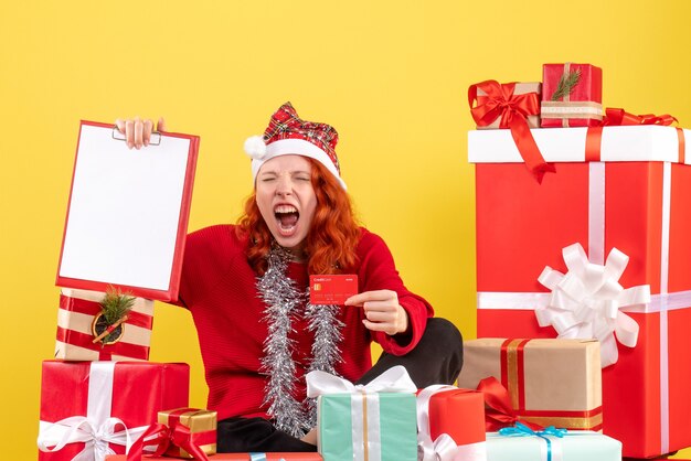 Vorderansicht der jungen Frau, die um Weihnachten sitzt, präsentiert Bankkarte auf gelber Wand