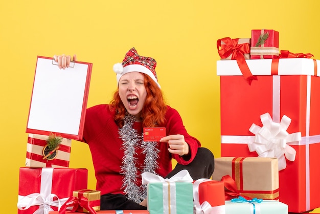 Vorderansicht der jungen Frau, die um Weihnachten sitzt, präsentiert Bankkarte auf gelber Wand