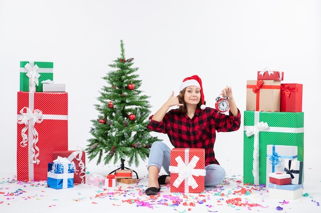 Vorderansicht der jungen Frau, die um Weihnachten sitzt, präsentiert das Halten von Uhren an der weißen Wand