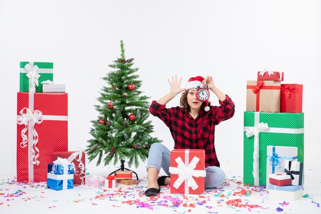 Vorderansicht der jungen Frau, die um Weihnachten sitzt, präsentiert das Halten von Uhren an der weißen Wand