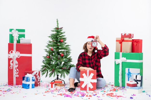 Vorderansicht der jungen Frau, die um Weihnachten sitzt, präsentiert das Halten von Uhren an der weißen Wand