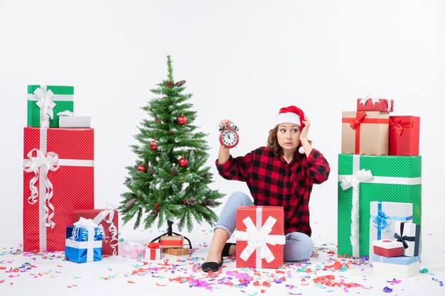 Vorderansicht der jungen Frau, die um Weihnachten sitzt, präsentiert das Halten von Uhren an der weißen Wand