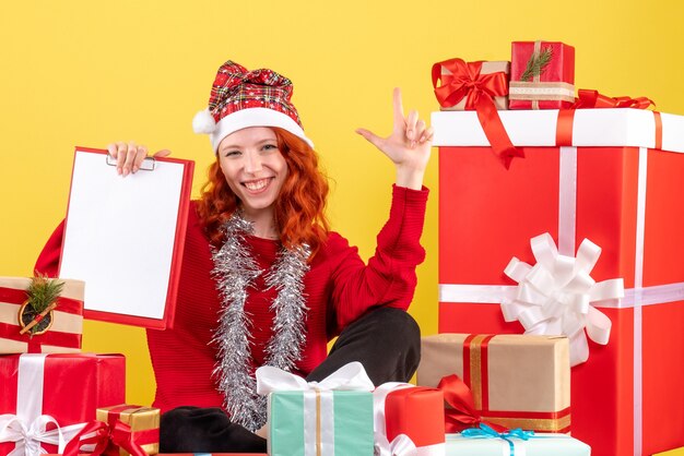 Vorderansicht der jungen Frau, die um Weihnachten sitzt, präsentiert mit Aktennotiz auf gelber Wand