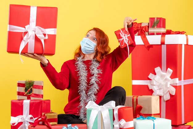 Vorderansicht der jungen Frau, die um Weihnachtsgeschenke in der Maske auf gelber Wand sitzt