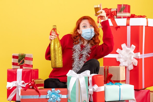 Vorderansicht der jungen Frau, die um Weihnachtsgeschenke in der Maske mit Champagner auf gelber Wand sitzt