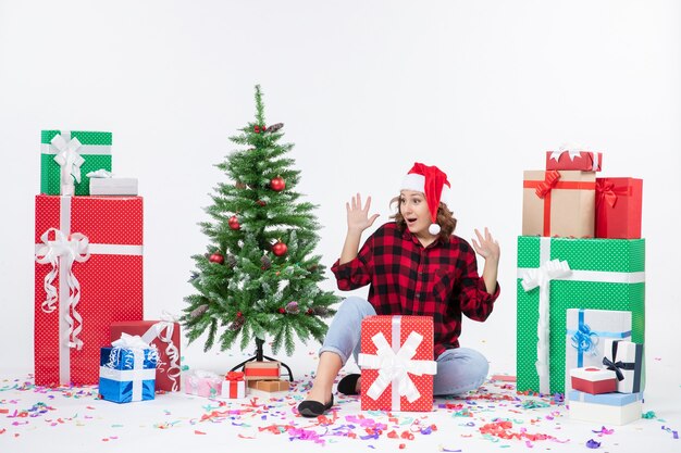 Vorderansicht der jungen Frau, die um Weihnachtsgeschenke und kleinen Feiertagsbaum auf weißer Wand sitzt