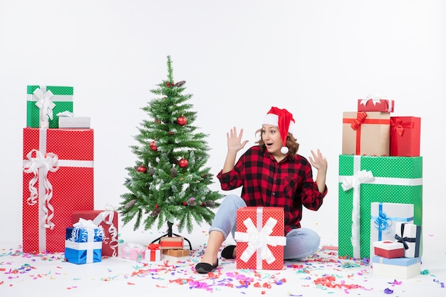 Vorderansicht der jungen Frau, die um Weihnachtsgeschenke und kleinen Feiertagsbaum auf weißer Wand sitzt