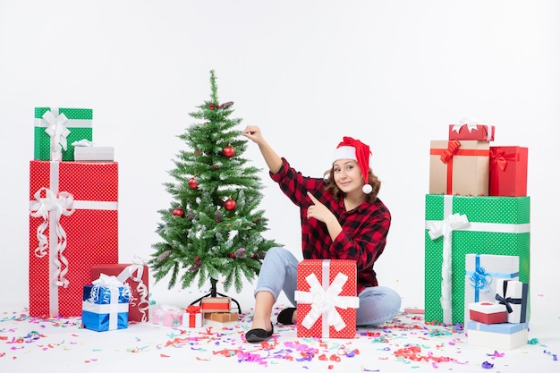 Vorderansicht der jungen Frau, die um Weihnachtsgeschenke und kleinen Feiertagsbaum auf weißer Wand sitzt