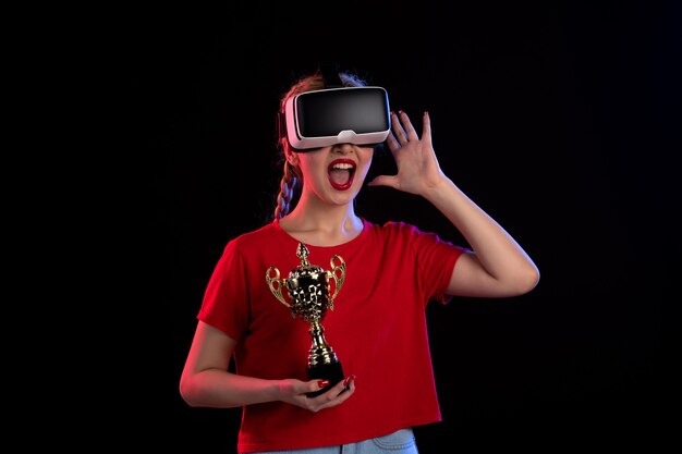 Vorderansicht der jungen Frau, die vr mit Siegerpokal an der dunklen Wand spielt