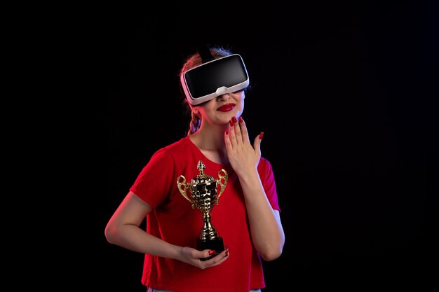 Vorderansicht der jungen Frau, die vr mit Siegerpokal an der dunklen Wand spielt