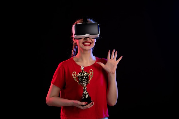 Vorderansicht der jungen Frau, die vr mit Siegerpokal auf dunklem Boden visuellem d-Tech-Spiel spielt