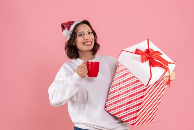 Vorderansicht der jungen Frau, die Weihnachtsgeschenk und Tasse Tee auf der rosa Wand hält