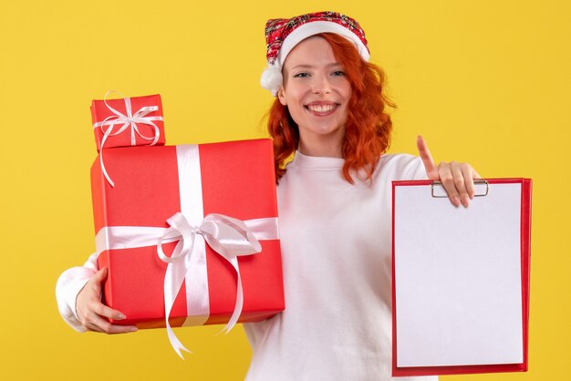 Kostenloses Foto vorderansicht der jungen frau, die weihnachtsgeschenke und aktennotiz auf gelber wand hält