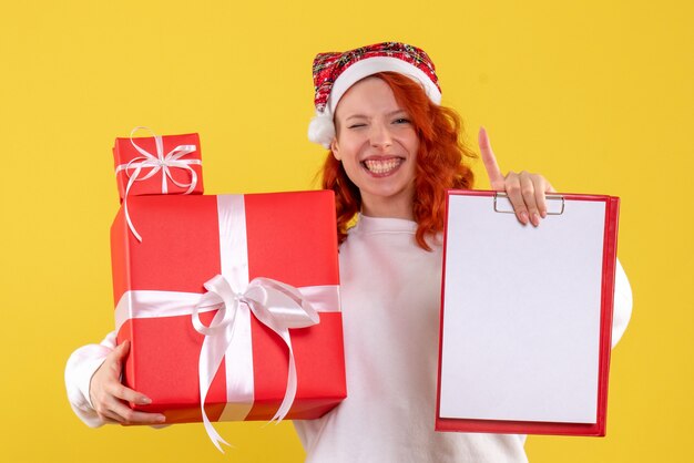 Kostenloses Foto vorderansicht der jungen frau, die weihnachtsgeschenke und aktennotiz auf gelber wand hält