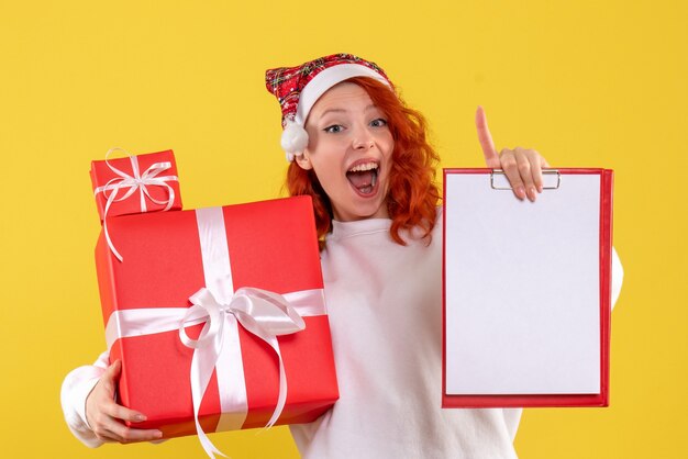 Vorderansicht der jungen Frau, die Weihnachtsgeschenke und Aktennotiz auf gelber Wand hält