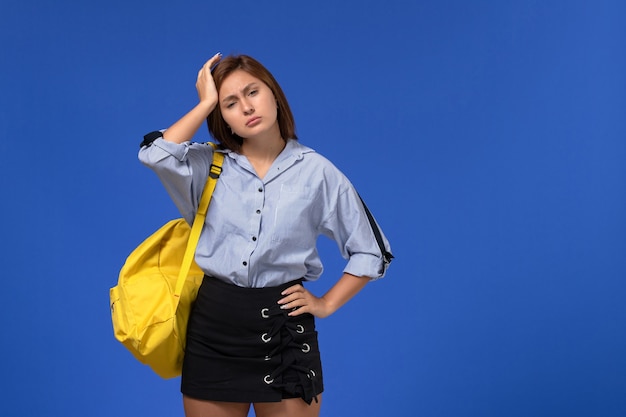 Vorderansicht der jungen Frau im blauen Hemd, das gelben Rucksack trägt, der Kopfschmerzen auf blauer Wand hat