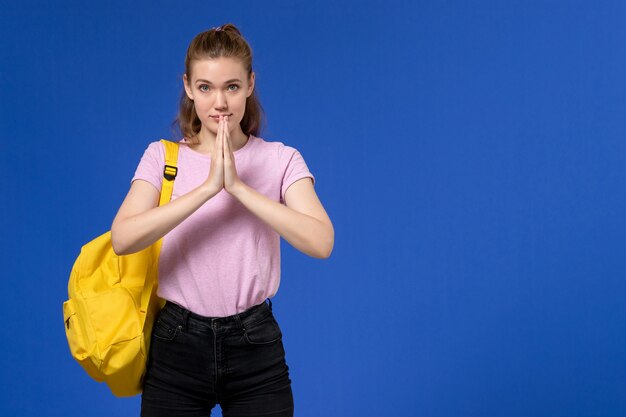 Vorderansicht der jungen Frau im rosa T-Shirt, das gelben Rucksack auf der hellblauen Wand trägt