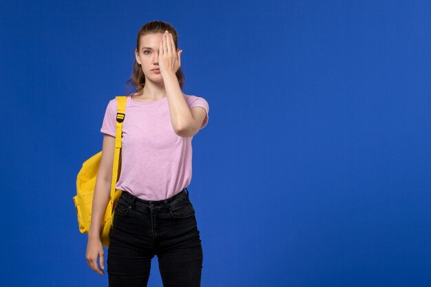 Vorderansicht der jungen Frau im rosa T-Shirt, das gelben Rucksack auf der hellblauen Wand trägt