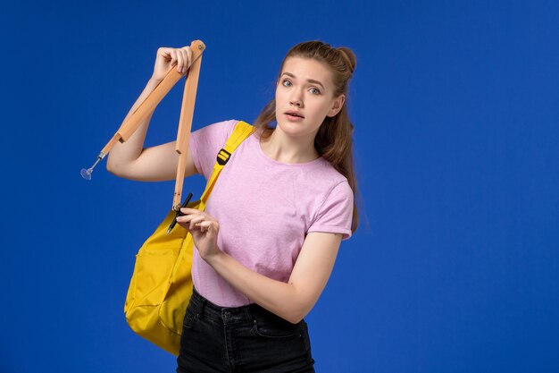 Vorderansicht der jungen Frau im rosa T-Shirt, das gelben Rucksack hält, der Holzfigur an der blauen Wand hält