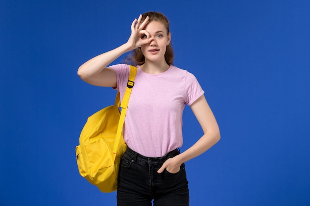 Vorderansicht der jungen Frau im rosa T-Shirt, das gelben Rucksack trägt, der auf der hellblauen Wand aufwirft