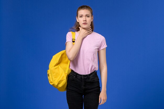 Vorderansicht der jungen Frau im rosa T-Shirt, das gelben Rucksack trägt, der Halsschmerzen an der blauen Wand hat