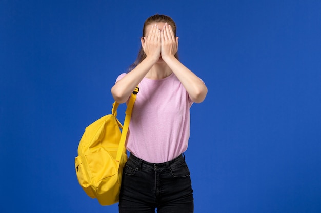 Vorderansicht der jungen Frau im rosa T-Shirt, das gelben Rucksack trägt, der ihr Gesicht auf der hellblauen Wand schließt