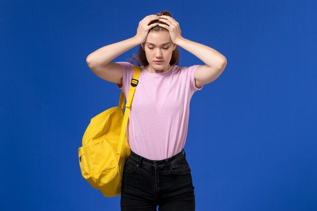Vorderansicht der jungen Frau im rosa T-Shirt, das gelben Rucksack trägt, der Kopfschmerzen auf hellblauer Wand hat
