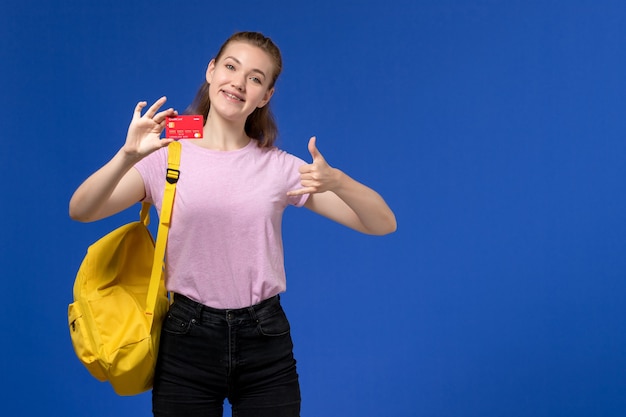 Vorderansicht der jungen Frau im rosa T-Shirt tragenden gelben Rucksack, der rote Plastikkarte mit Lächeln auf der blauen Wand hält