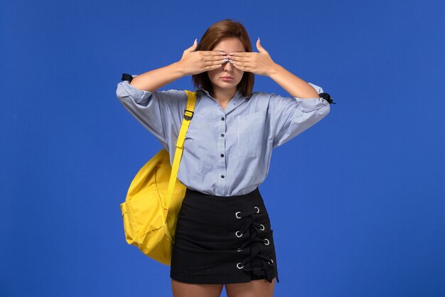 Vorderansicht der jungen Frau im schwarzen Rock des blauen Hemdes, der gelben Rucksack trägt, der ihre Augen auf hellblauer Wand schließt