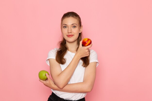 Vorderansicht der jungen Frau im weißen T-Shirt, das frischen grünen Apfel mit Pfirsich lächelnd auf hellrosa Wand hält
