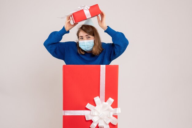 Vorderansicht der jungen Frau in der Geschenkbox in Maske mit Geschenken auf weißer Wand