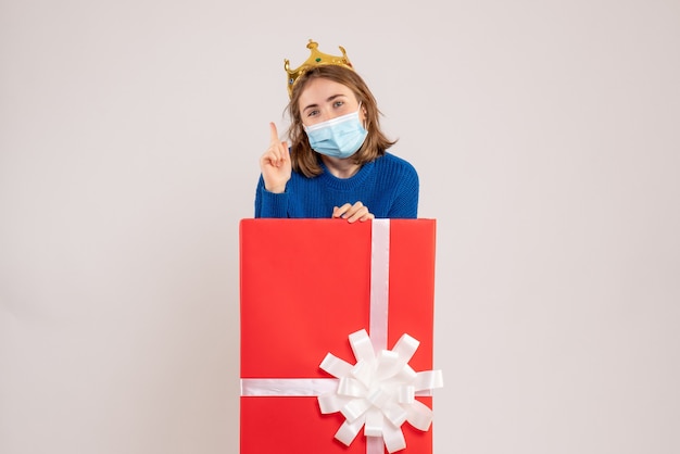 Vorderansicht der jungen Frau in der Geschenkbox in steriler Maske auf weißer Wand